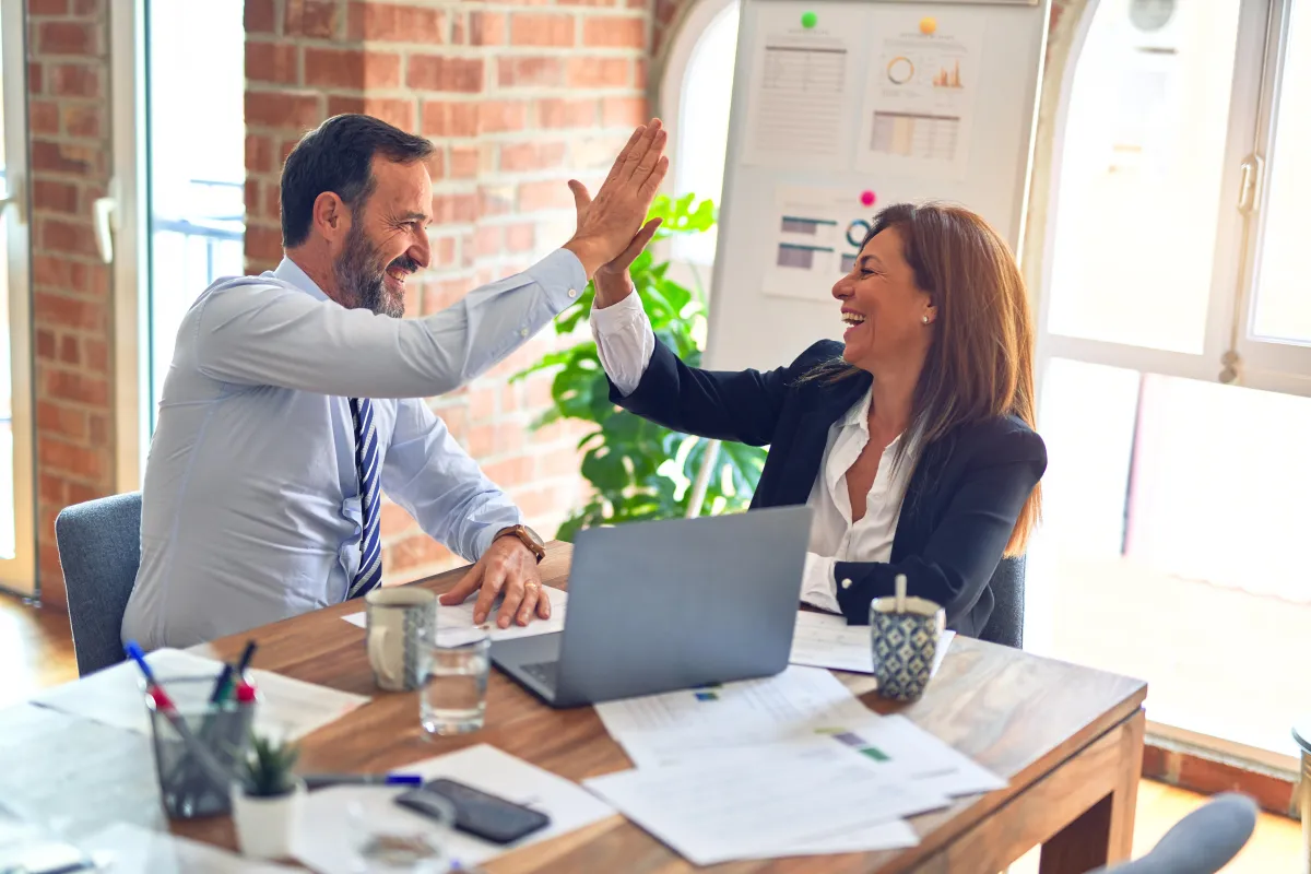 Coworkers sitting at table with laptops are high-fiving in celebration