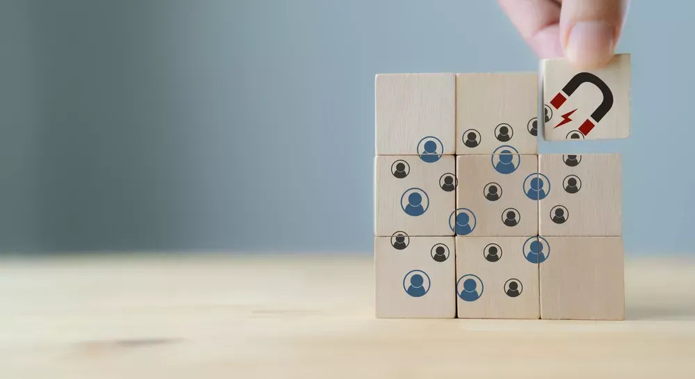 A hand places a wooden block with a magnet icon atop a stack of blocks, symbolizing Sales & Marketing. Other blocks, illustrated with blue and black human figures, are arranged in a 4x3 grid on a light wooden surface in front of a plain background.