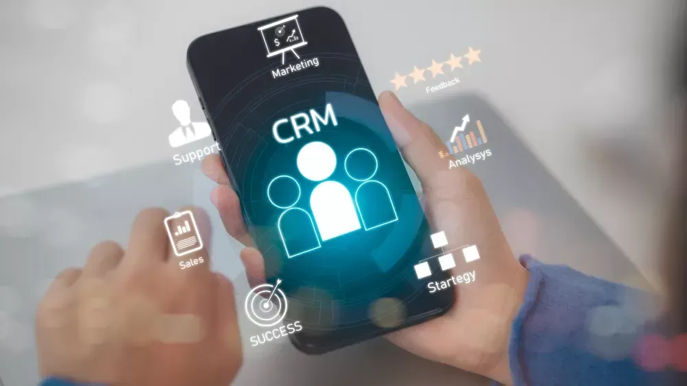 A person holds a smartphone showcasing a CRM icon, surrounded by terms like Marketing, Feedback, Sales, Support, Analysis, and Strategy. Digital graphics envelop the phone, reflecting business and technology concepts tied to CRM innovation.