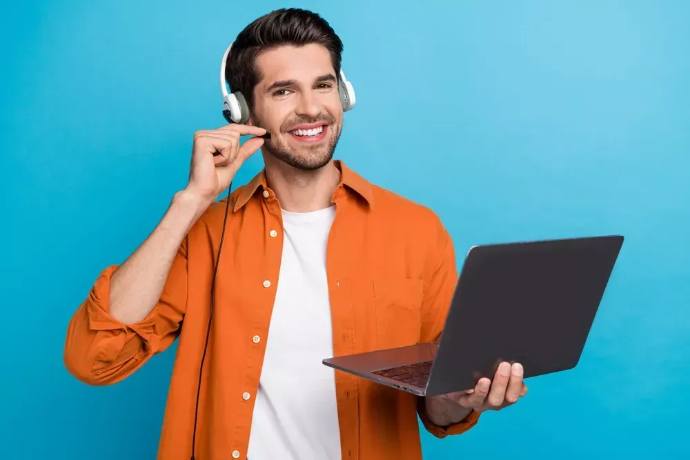 A smiling man wearing headphones and an orange shirt holds a laptop open in one hand, offering virtual assistance while gesturing with the other, set against a solid blue background