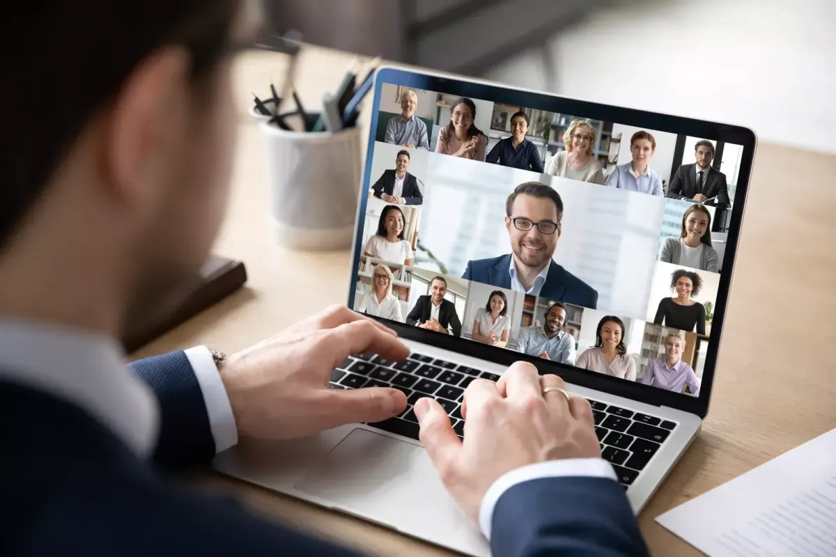 A person participates in a video conference call on a laptop with multiple participants visible in individual windows on the screen. The meeting takes place in a well-lit room, with a pen holder and notepad nearby, as they discuss CRM strategies guided by their virtual assistant..