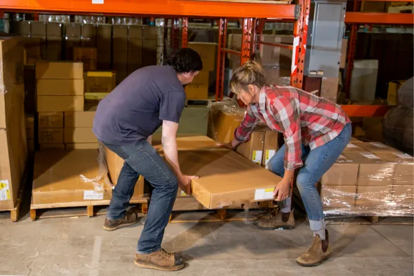 An industrial warehouse, workplace safety topic. Two employees correctly lifting a heavy box together.