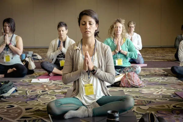 Group of adults practicing seated meditation in a yoga class, with eyes closed and hands in prayer pose.
