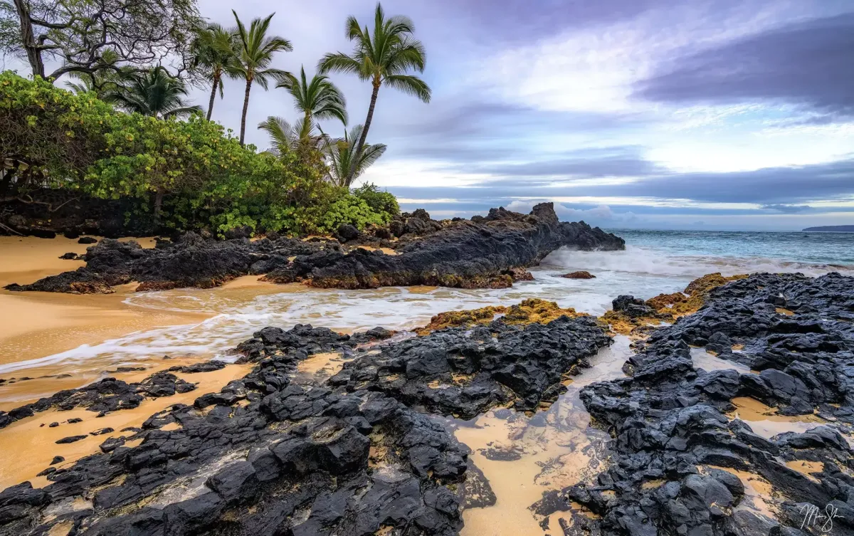 Large coastal rocks holding steady as waves crash along a tropical shoreline.