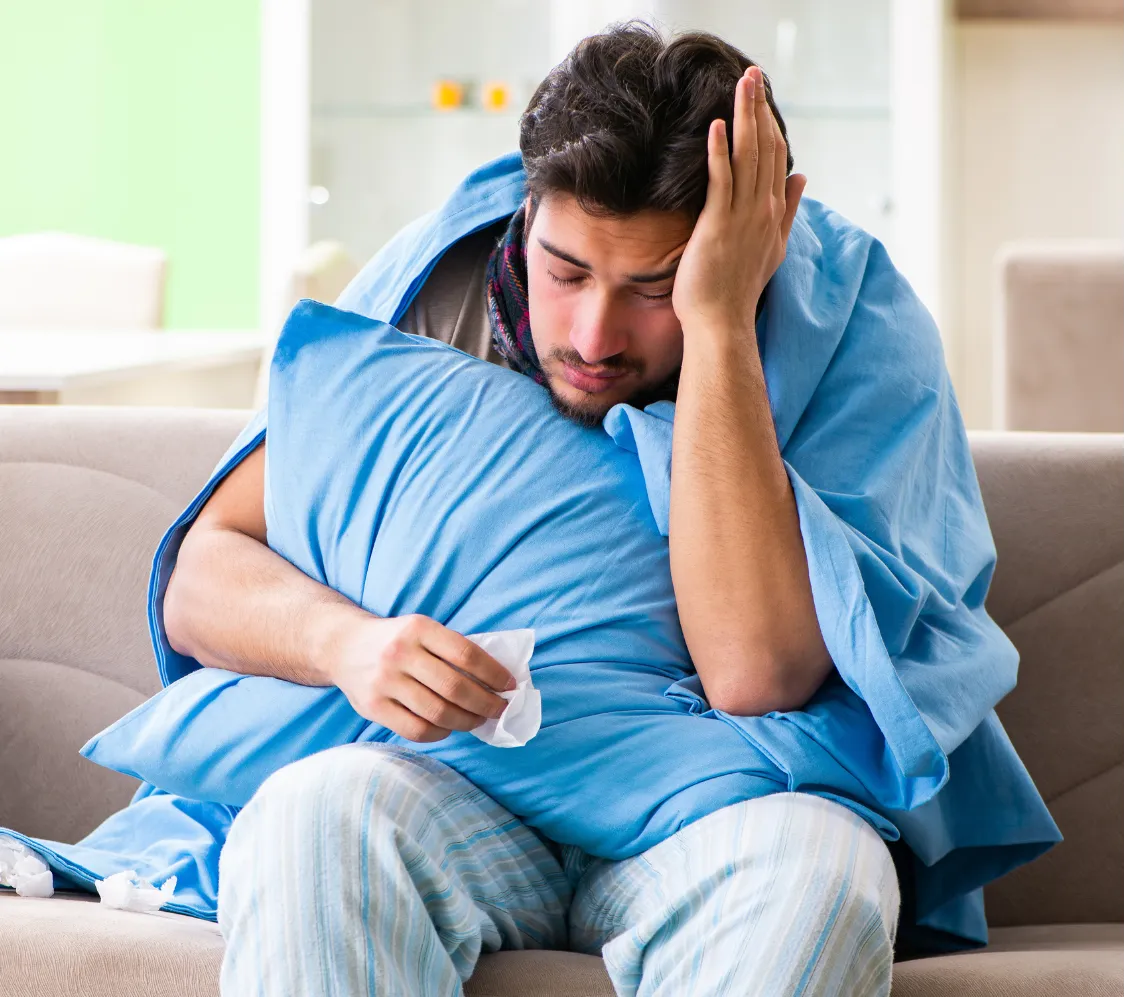 Man sitting on a couch surrounded by tissues, pillows, and blankets, depicting illness and fatigue.