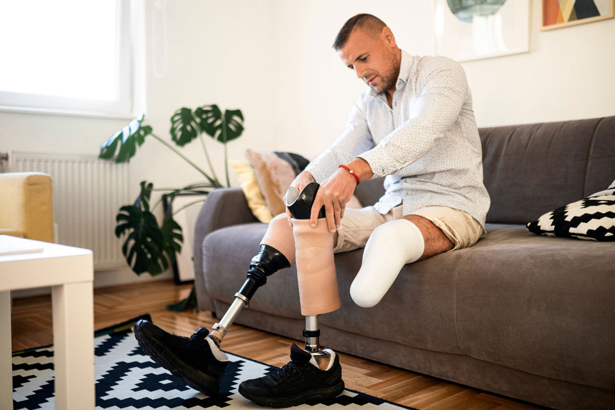 Male patient sits on a couch adjusting his below-knee prosthetic leg, demonstrating daily prosthetic care.