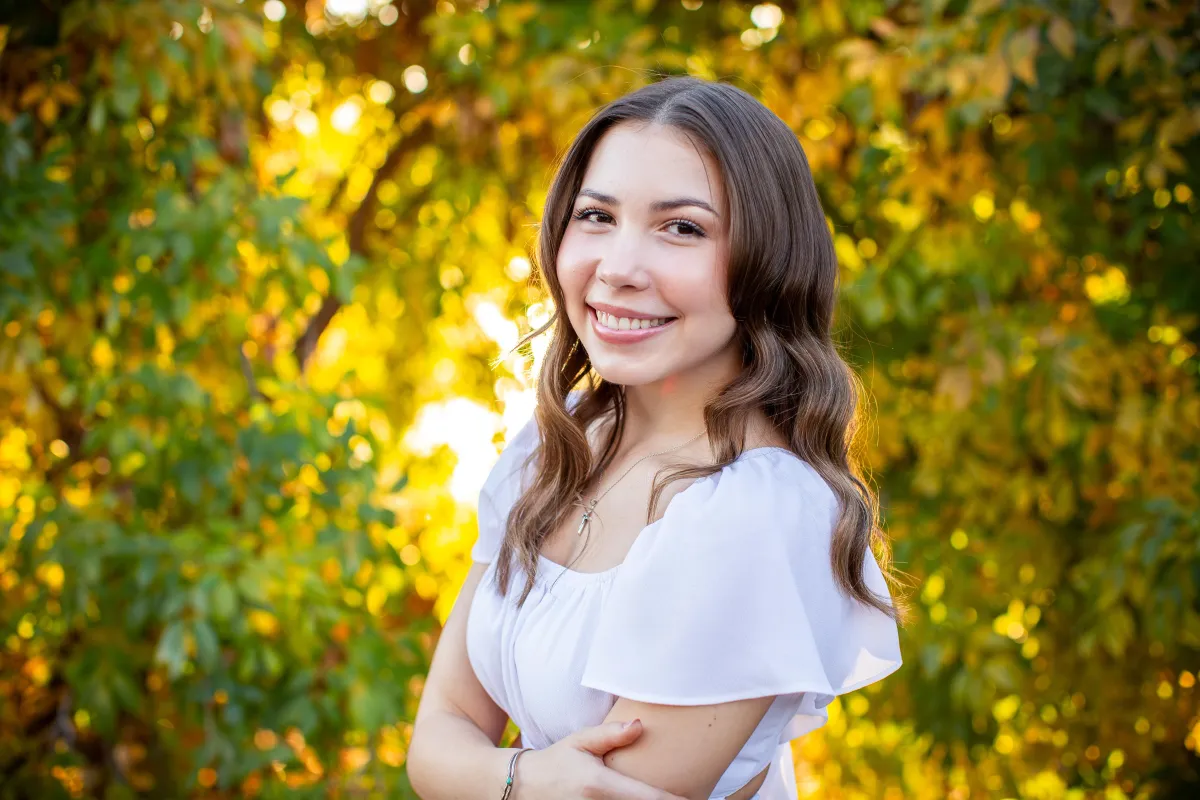 Hispanic high school senior girl smiling during a fall portrait session at the Riparian Preserve in Gilbert, AZ, surrounded by golden autumn foliage — taken by Andria May Photography.