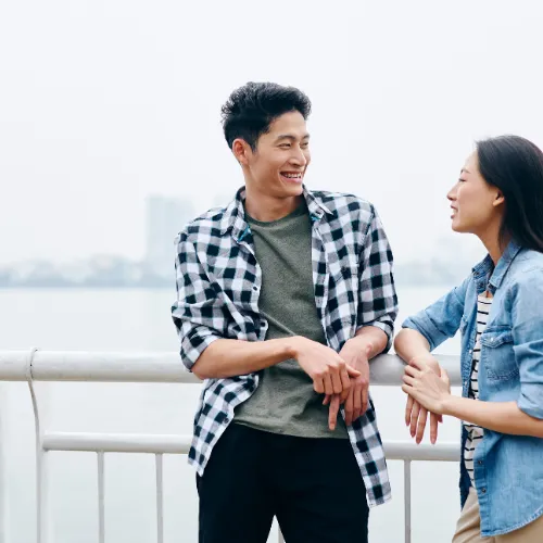 A young man and woman are standing by a waterfront railing, engaged in a cheerful conversation. The man, wearing a green t-shirt and a black-and-white checkered shirt, is leaning against the railing, smiling at the woman. The woman, dressed in a denim jacket and a striped shirt, is gesturing with her hands as she talks to him. The background is a hazy view of a city skyline across the water.