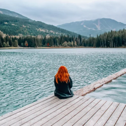 A person with long red hair sits at the edge of a wooden dock, facing a serene lake surrounded by dense forest and mountains. The scene is tranquil, with the individual appearing to be in a moment of contemplation or relaxation, enjoying the natural beauty and calm waters. The sky is overcast, adding a peaceful and reflective mood to the setting.