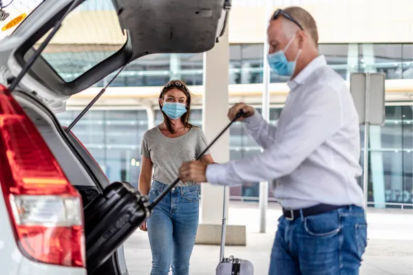 A man and a woman, both wearing face masks, are at an airport. The man, dressed in a white shirt and jeans, is placing a suitcase into the trunk of a car. The woman, wearing a gray t-shirt and jeans, stands nearby watching. The background shows the exterior of an airport terminal with large windows.