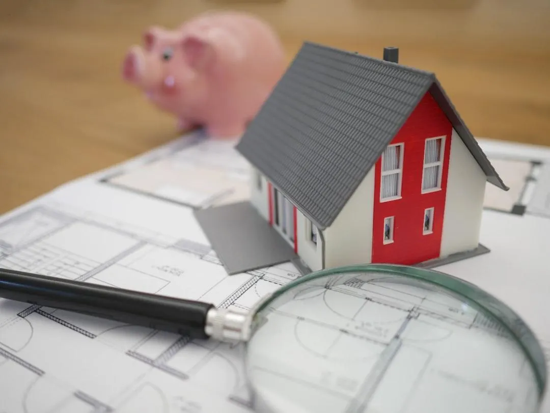 This image features a small model house placed on top of architectural blueprints, accompanied by a magnifying glass and a pen, symbolizing careful planning and analysis in the home buying or mortgage process. In the background, a pink piggy bank represents financial savings, highlighting the importance of budgeting and financial preparedness when purchasing a home.