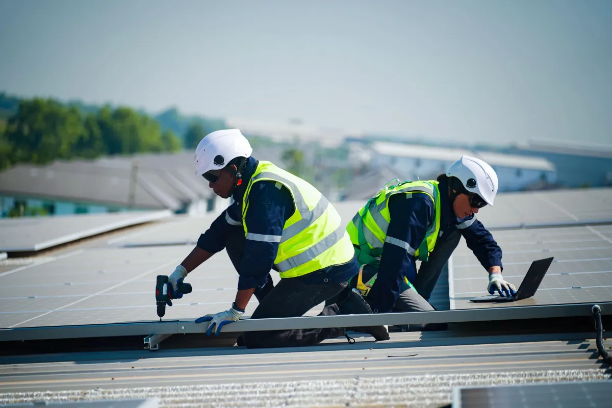 Two technicians in hard hats and safety vests installing solar panels on a large, light-colored commercial roof. 