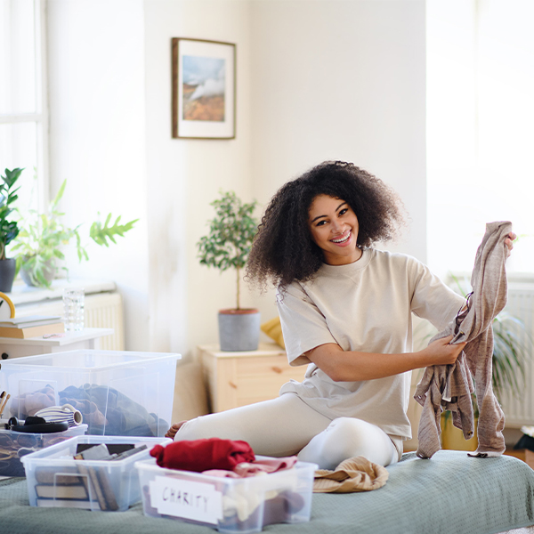 A smiling woman with curly hair is sitting on a bed, happily folding clothes and sorting items into clear storage bins, one labeled "CHARITY.
