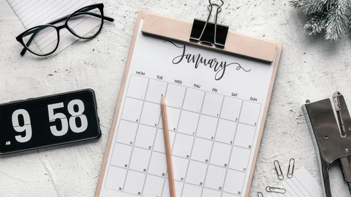 A desk with a calendar, glasses, a clock, and a stapler.
