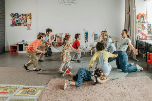 Children and two adults play and dance in a brightly lit classroom. The room is decorated with colorful art and toys. Some children hold balloons while others move energetically. The atmosphere is playful and lively.