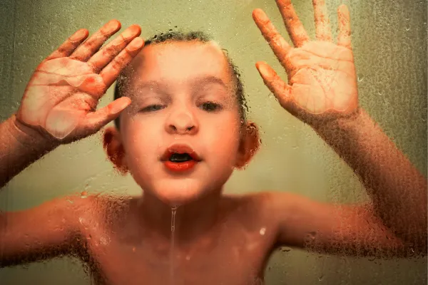 A young child with short hair presses their face and hands against a wet glass surface. Water droplets are visible on the glass, and the background is softly blurred, giving a playful and innocent atmosphere.