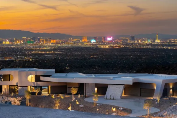 A modern, luxurious mansion with sleek white architecture sits on a hillside in Henderson, NV, overlooking the vibrant Las Vegas skyline at sunset. The sky glows with warm hues of orange and pink, while the city lights sparkle below, framed by distant mountains. The foreground features minimalist landscaping with small trees and soft lighting, highlighting the property's elegance.