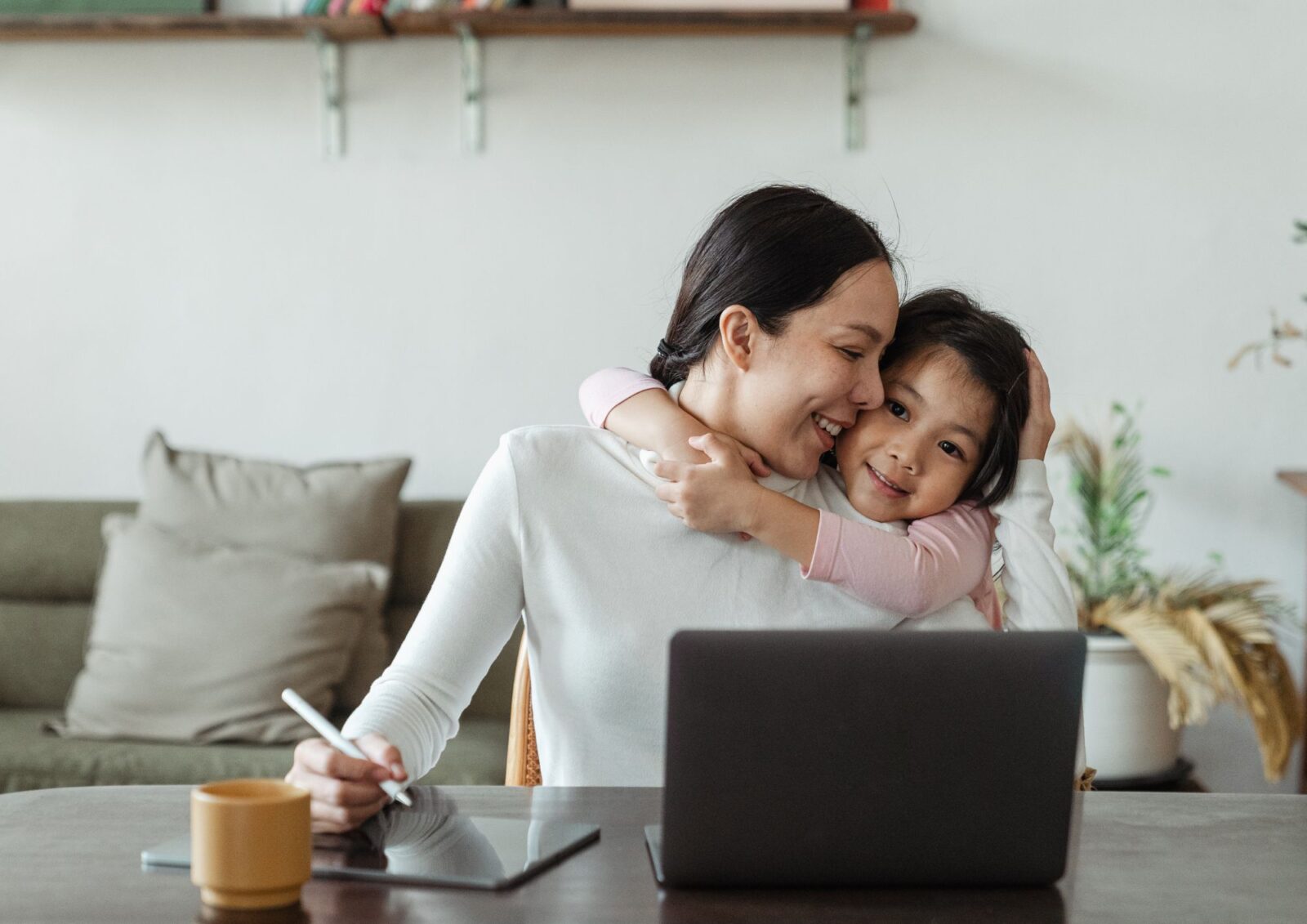 A smiling woman working on a laptop and tablet at a home desk while being happily hugged from behind by her young daughter.
