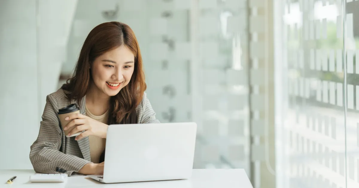 A smiling young businesswoman sitting at a desk, looking at her laptop screen while holding a takeaway coffee cup.