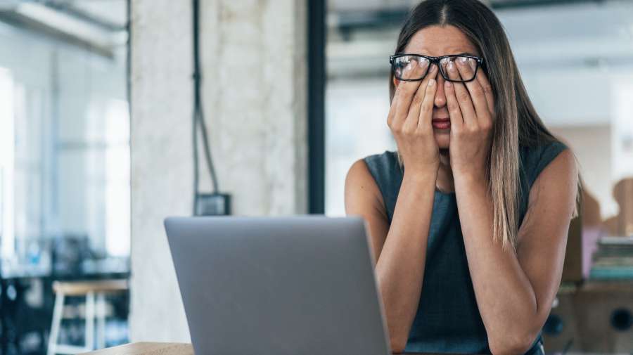 A woman with glasses is sitting at a desk in front of a laptop and covering her face with her hands in a gesture of stress or exhaustion.