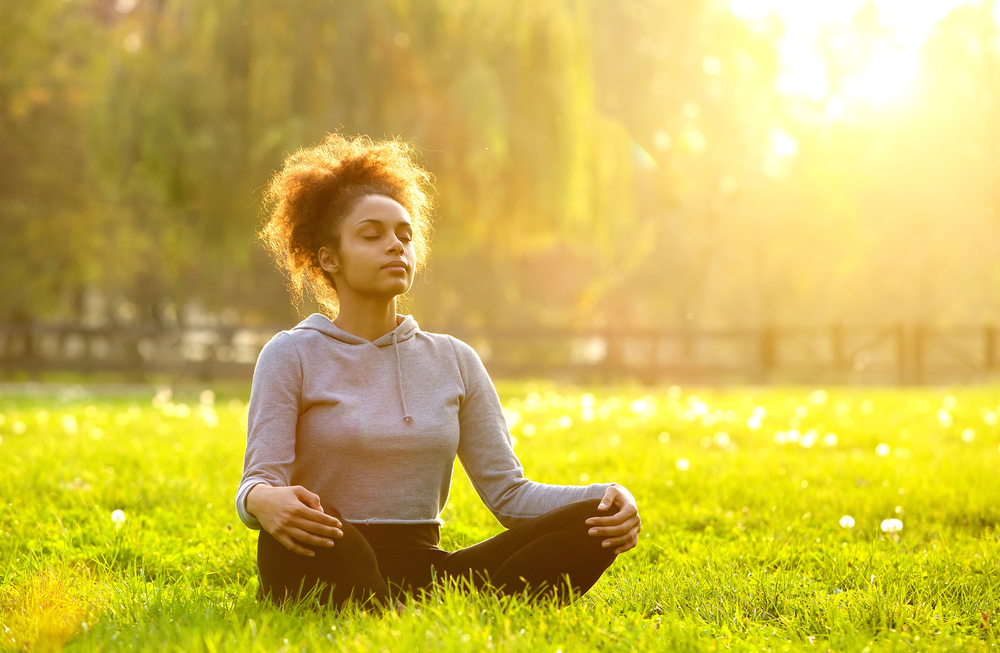 A young woman with curly hair is sitting cross-legged and meditating in a bright, sunny, grassy field.