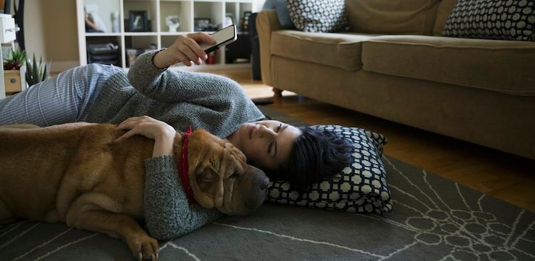 A woman is lying on the floor with a large, wrinkly brown dog resting its head on her arm, and she is looking at her smartphone.