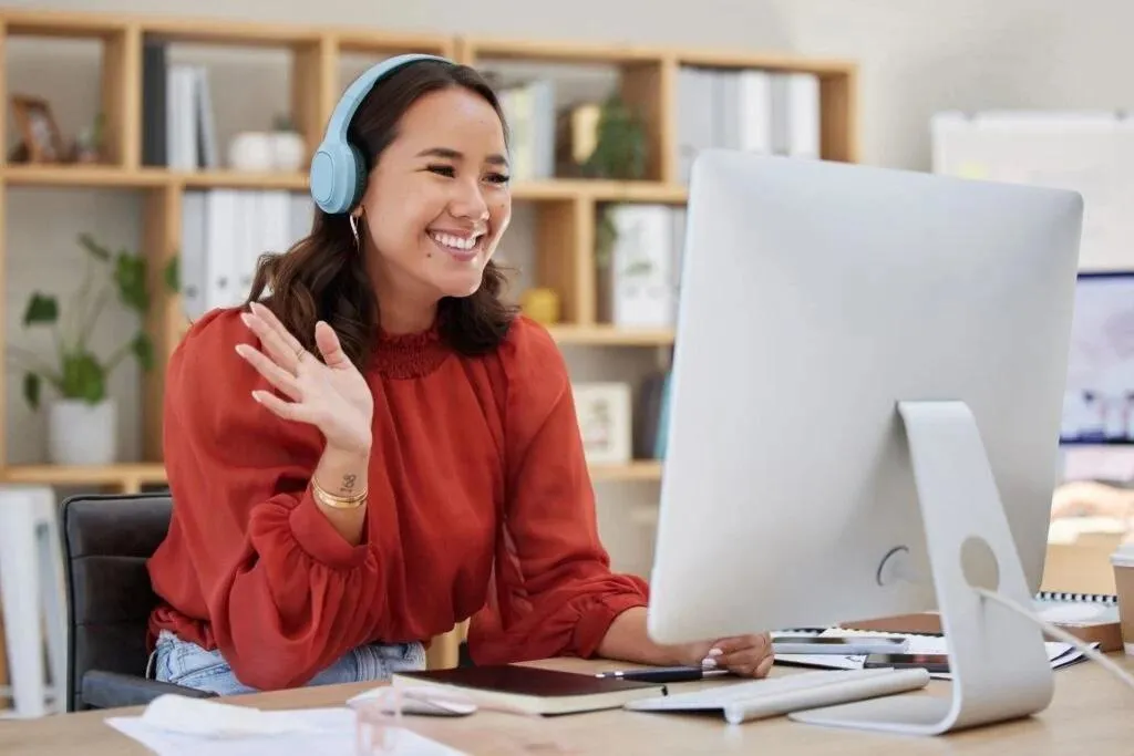 A smiling woman wearing blue over-ear headphones waves at a computer screen while seated at a desk in an office setting.