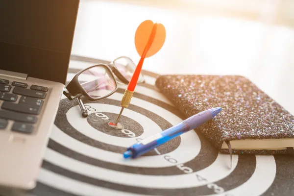 Dart in the middle of a target near a computer, journal, and writing pen.