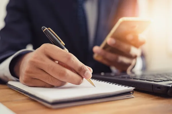 Person looking at their phone while taking notes on a piece of paper – image represents a marketing checklist for a business.
