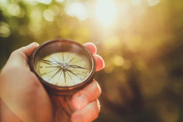 Hand holding an old-fashioned compass in the air as the warm sun illuminates the trees in the background – image represents branding guides. 