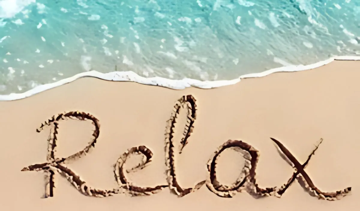 The word “Relax” written in soft sand at the edge of the ocean, with gentle turquoise waves approaching the shore.