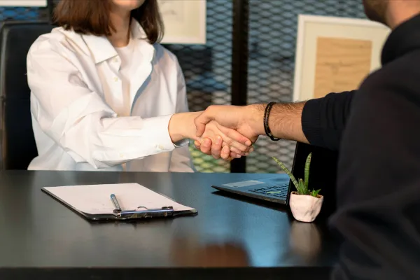 A man and woman greet each other with a handshake at a desk.