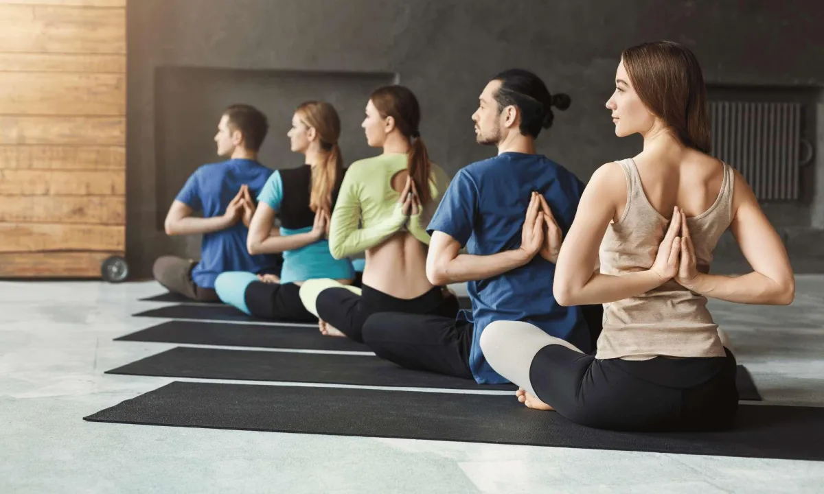 A group of people in a yoga class sit on mats, practicing a seated pose with hands