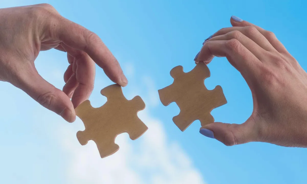 Two hands each holding a piece of a jigsaw puzzle against a blue sky background.