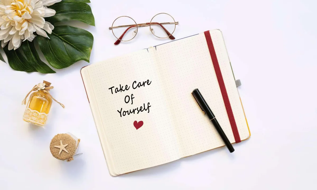 Notebook with "Take Care Of Yourself" message, glasses, pen, bottle, and flower.