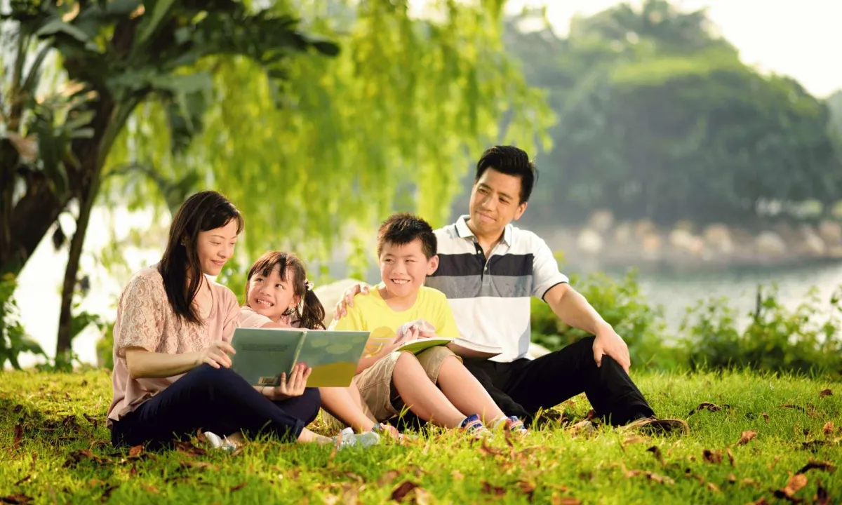 A family of four sits on the grass in a park, reading a book together.