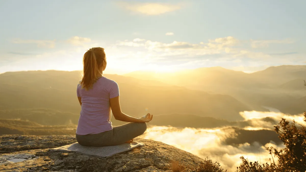 A person sitting on a rock, meditating at sunrise with mountains and clouds in the background.