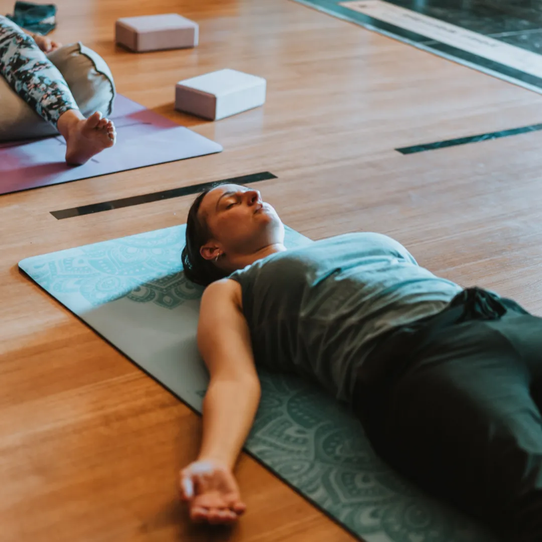 Practitioner in Savasana with legs elevated on a bolster, lying on the back with arms relaxed and eyes closed, supporting deep rest and gentle circulation.