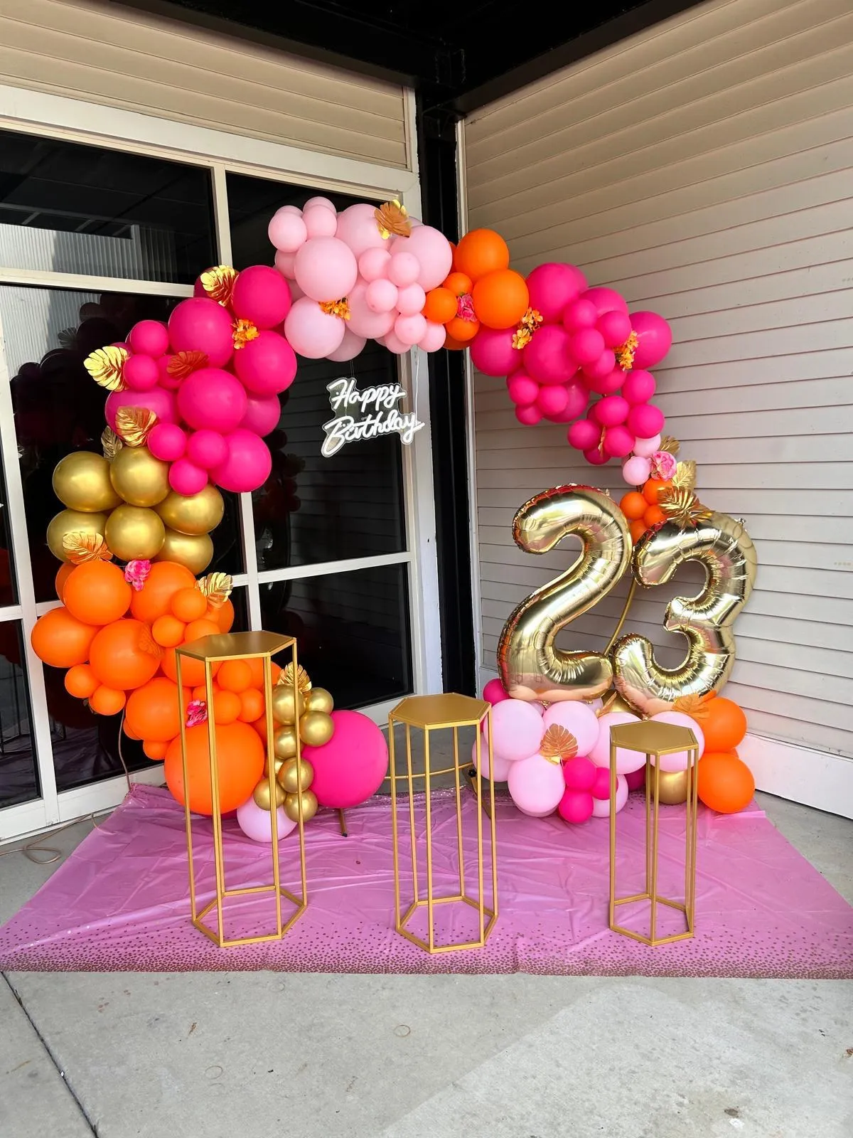 Vibrant pink, orange, and gold balloon arch with number 23 balloons for a birthday celebration, featuring floral accents and a ‘Happy Birthday’ sign