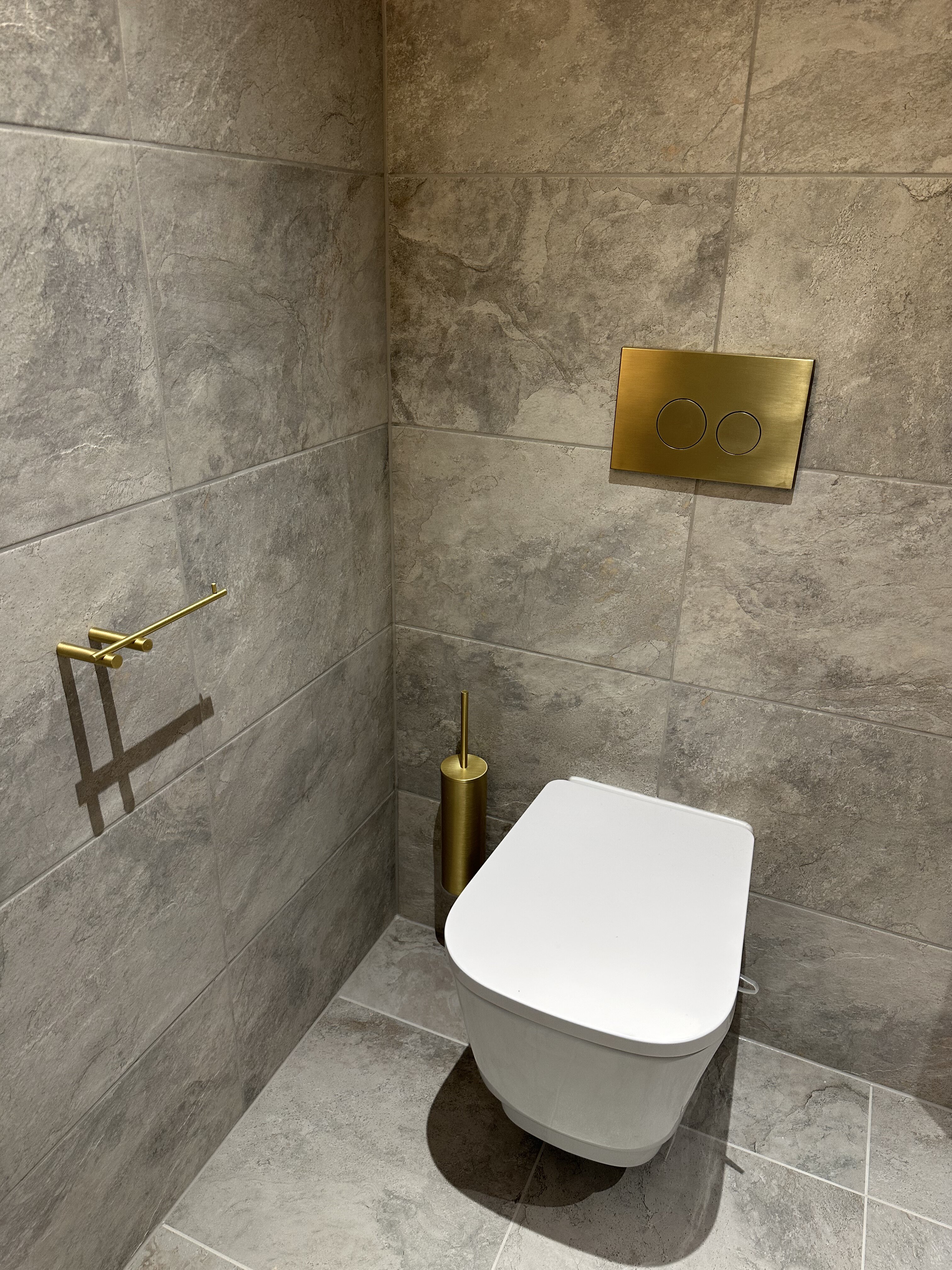 Luxury bathroom upgrade Glasgow; PJTC fitted premium brushed gold accessories, including a gold flush plate and towel bar, against textured grey stone-effect wall tiling.