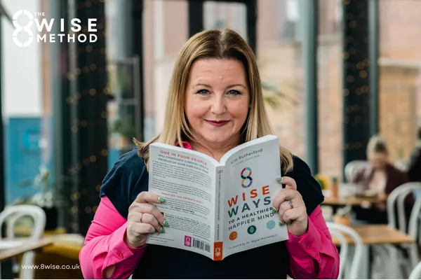 Kim Rutherford looking at camera holding a copy of her book 8 Wise Way to a Happier Healthier Mind