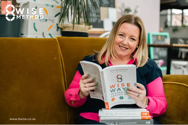 Kim Rutherford smiling to the camera holding a copy of her book 8 Wise Ways to a Happier Healthier Mind