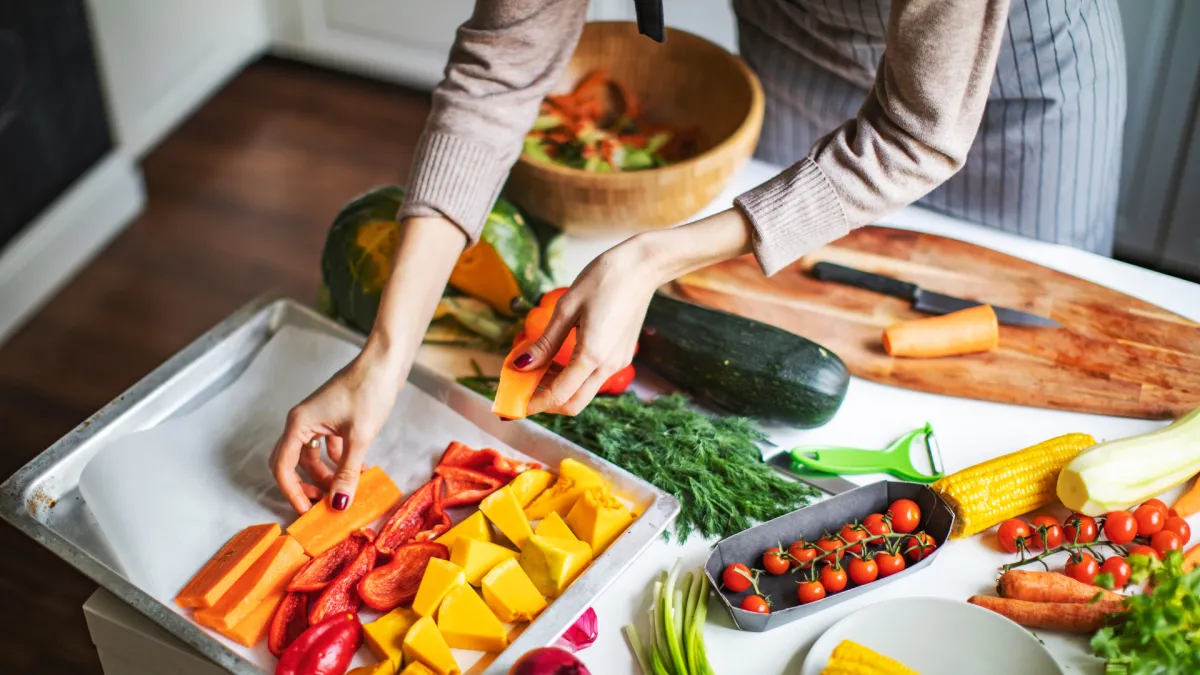 A person preparing a colorful variety of fresh vegetables in a bright kitchen. They are arranging sliced squash, red bell peppers, and carrots on a baking tray lined with parchment paper, surrounded by zucchini, corn, tomatoes, and herbs—representing healthy meal prep and whole-food cooking.