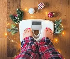 A person in red plaid pajamas standing on a bathroom scale, surrounded by festive holiday decorations and string lights, representing holiday weight tracking, seasonal health awareness, or staying accountable during celebrations.