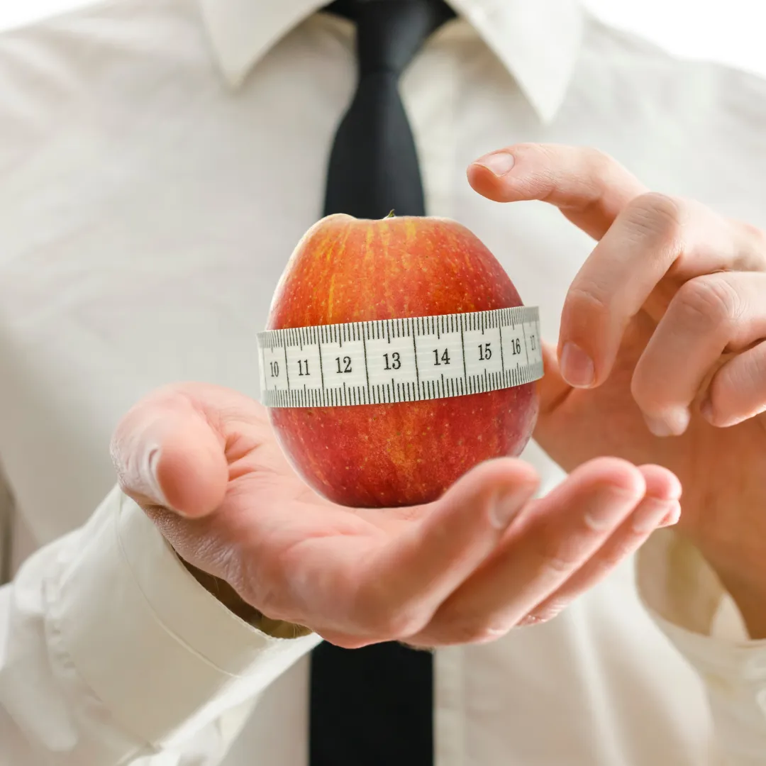 A person in a white shirt and black tie holds a red apple wrapped with a measuring tape, symbolizing health, nutrition, or weight loss. The focus is on the apple and tape, suggesting diet or portion control.