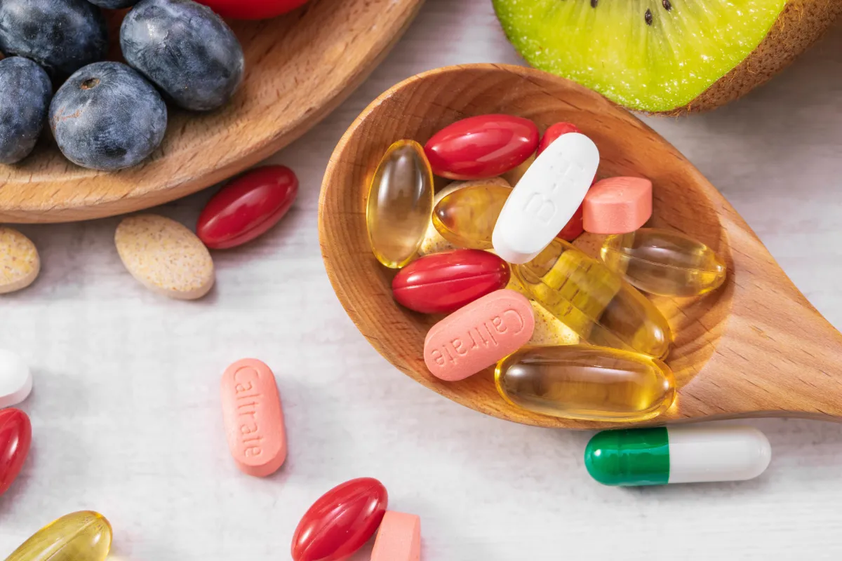 Assorted vitamins and supplements in a wooden spoon beside fresh fruits, representing the balance between whole foods and targeted supplementation during Hyper-Ketosis.