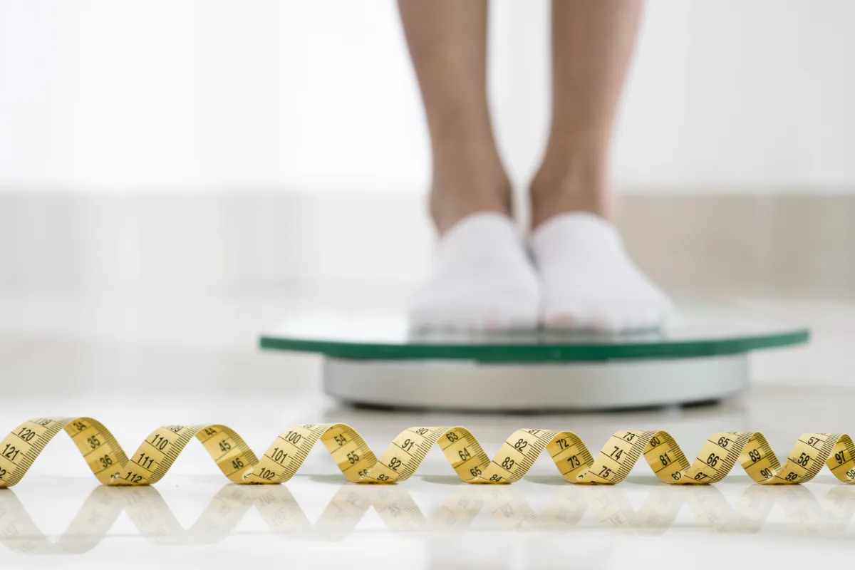 Person standing on a glass scale with a yellow measuring tape in the foreground, symbolizing tracking progress and measuring body changes during Hyper-Ketosis.