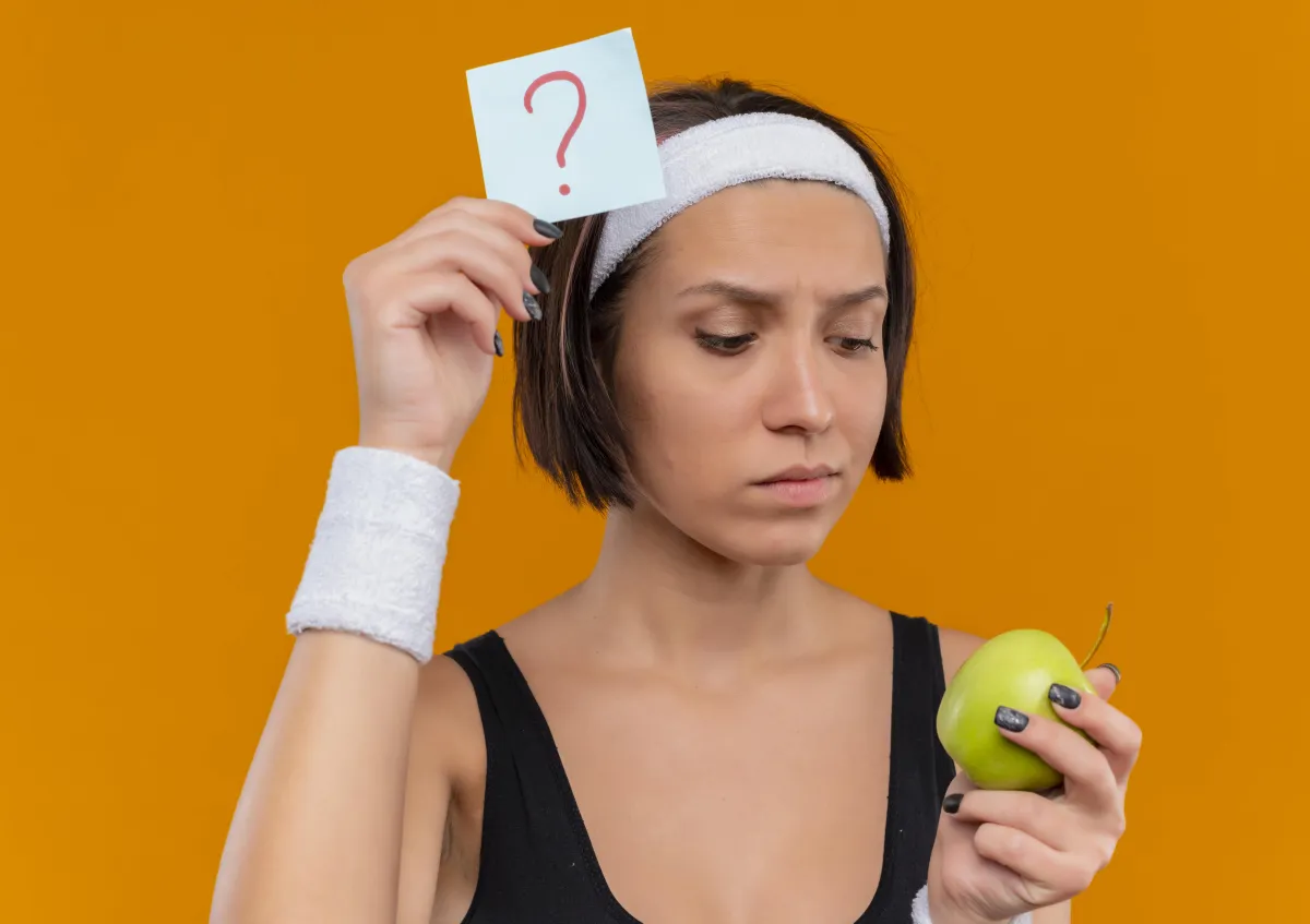 Woman in workout gear holding a green apple and question mark note, symbolizing common questions about Hyper-Ketosis nutrition.