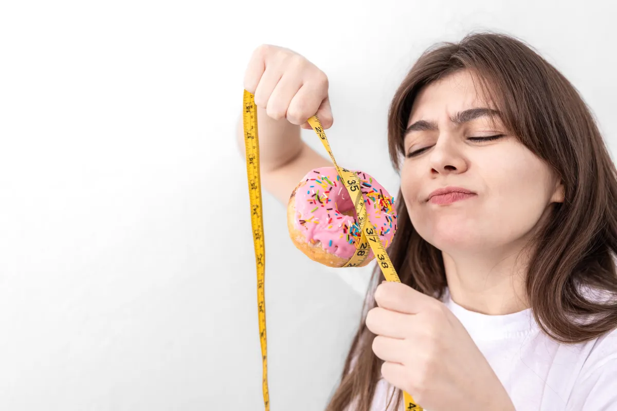 Young woman measuring a pink frosted donut with a tape measure, showing the mental struggle between food temptation and Hyper-Ketosis goals.