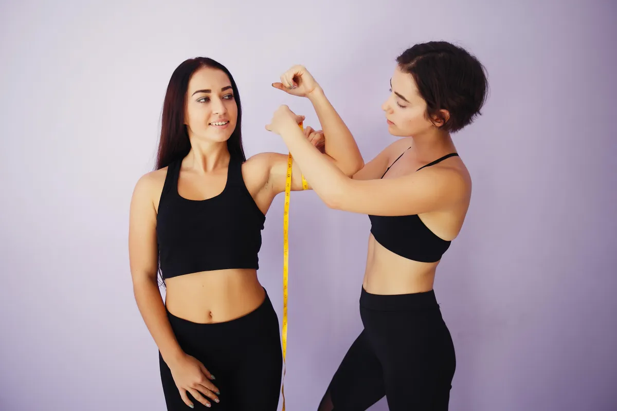 Two women in workout clothes measuring arm progress together, representing accountability and support in a Hyper-Ketosis fitness journey.